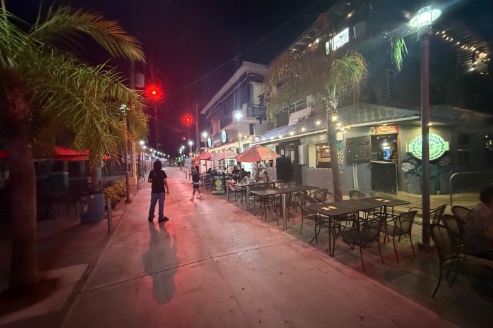 Night scene of a lively street with outdoor dining and palm trees.
