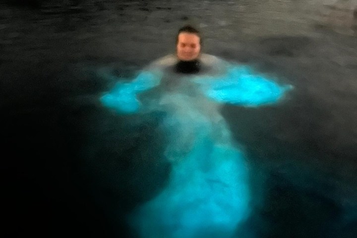 Person swimming in bioluminescent water at night with glowing blue light.